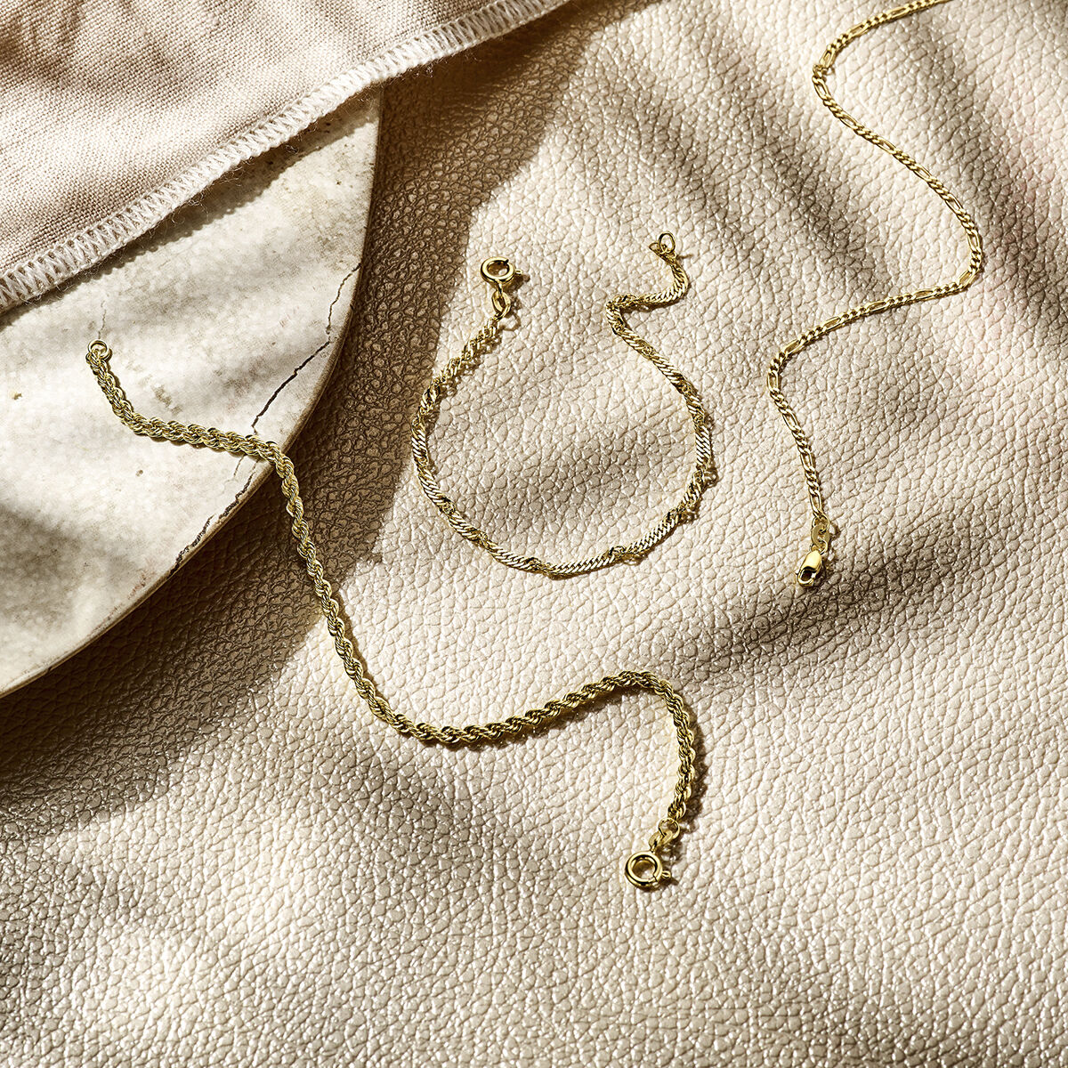 Drie delicate 14K geelgouden sieraden, waaronder de Van der Kooij Collectie armband figaro 1,8 mm, liggen op een beige oppervlak met zachte schaduwen en een stukje lichte stof zichtbaar in de hoek.