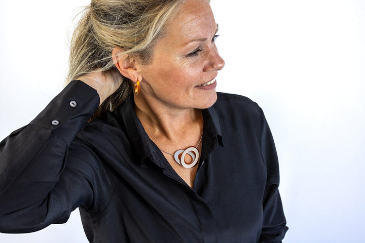 Een blonde vrouw met haar haar naar achteren gebonden glimlacht terwijl ze wegkijkt. Ze draagt een elegant zwart overhemd met knopen, oranje oorringen en een CLIC Creations Collier Ovalen op elkaar C70 ketting met in elkaar grijpende hangers. Haar hand raakt zachtjes haar nek aan tegen een effen witte achtergrond.