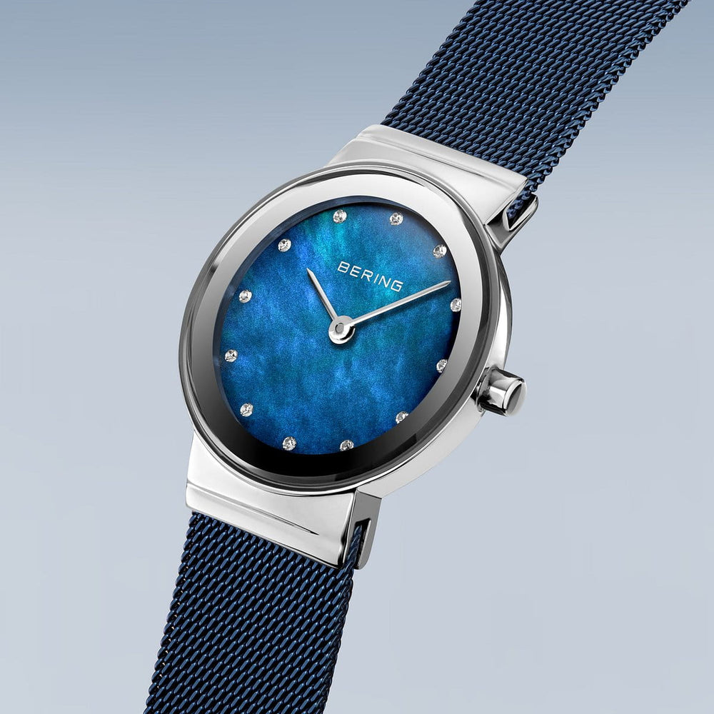 Het Bering dameshorloge 10126-307 heeft een zilverkleurige kast, een opvallende blauwe wijzerplaat met ronde markeringen, een bijpassende blauwe mesh band, saffierglas kristal en quartz uurwerk tegen een zachtverloop achtergrond.