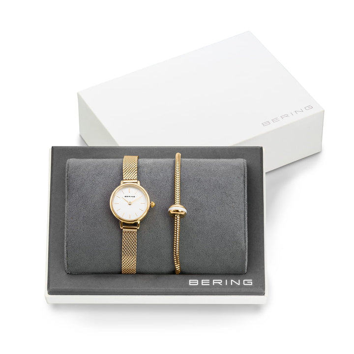 De Bering 11022-334-Lovely-1-GWP190 set bestaat uit een gouden dameshorloge met saffierglas, quartz uurwerk en witte wijzerplaat, gecombineerd met een eenvoudige gouden armband. Het horloge wordt weergegeven in een open doos met een grijze binnenkant en het Bering-logo op het deksel, en pronkt met een meshband.