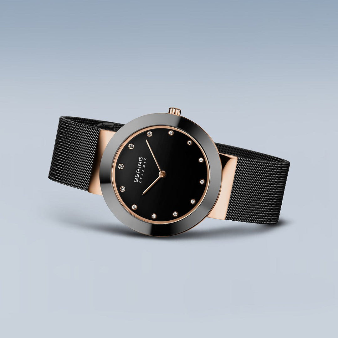 Het Bering 11435-166 polshorloge heeft een zwarte wijzerplaat met gouden markeringen, een gouden lunette en een minimalistisch design. Het omvat een zwarte mesh band, Quartz-uurwerk uurwerk, duurzaam Saffierglas en wordt elegant gepresenteerd tegen een zachtblauwe achtergrond.