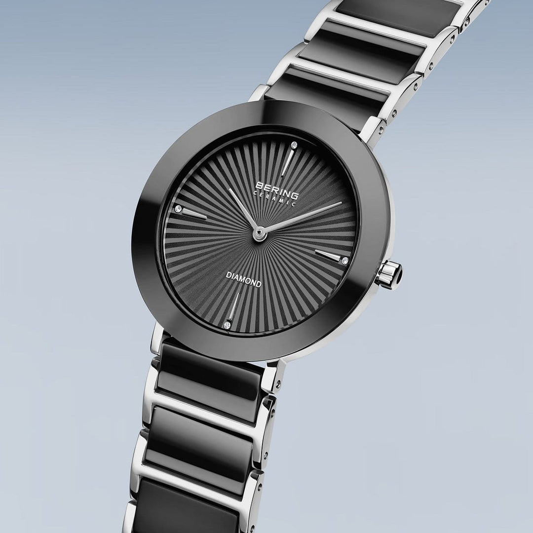 De Bering 11435-702 dameshorloge heeft een ronde zwarte wijzerplaat met zilveren wijzers, radiale patronen en "Diamond" onderaan. Het minimalistische ontwerp wordt aangevuld door een zwarte band met zilveren schakels voor een luxe uitstraling.