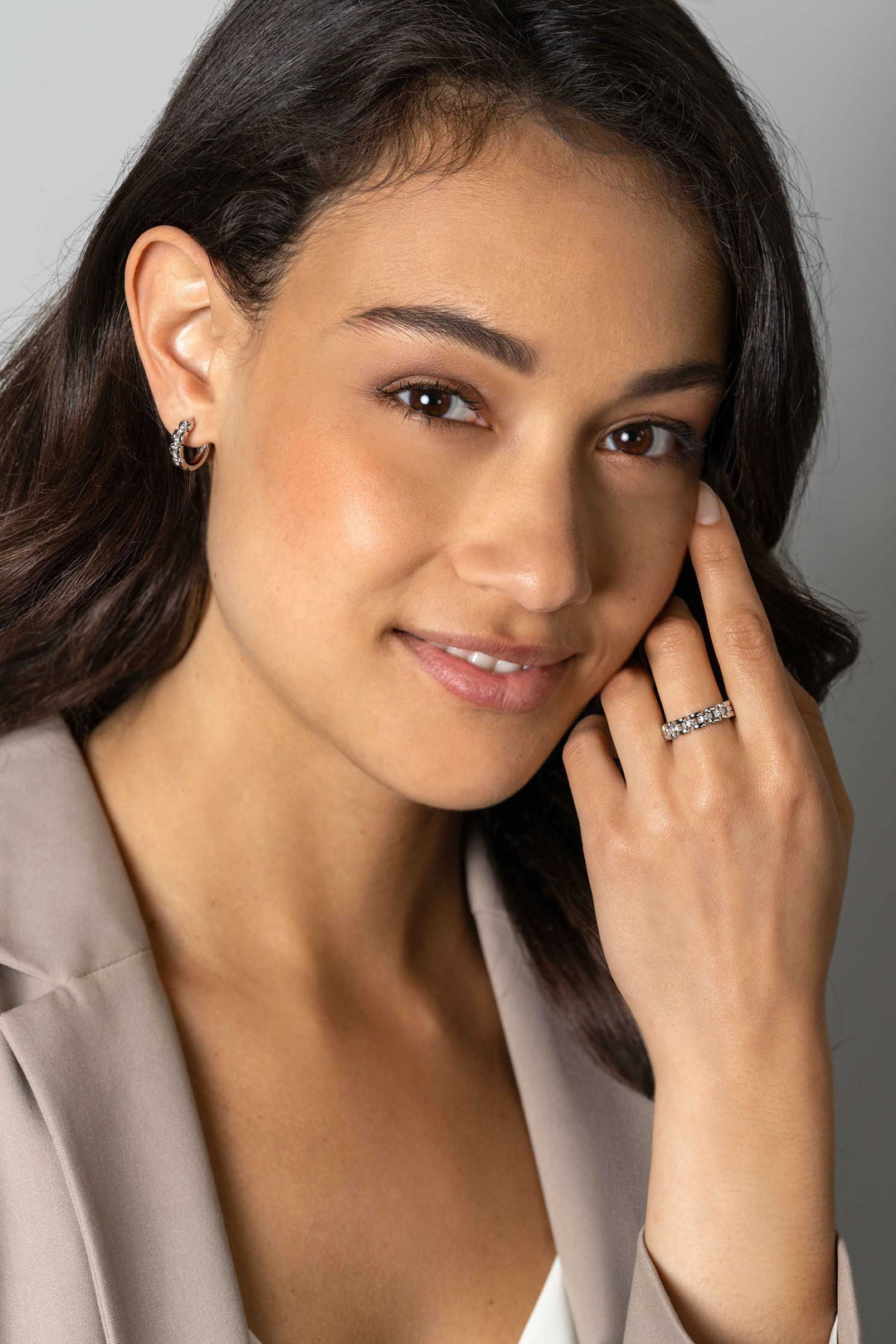 Een vrouw met golvend donker haar glimlacht zachtjes in een beige blazer, pronkt met een zilveren ring en draagt Zinzi Oorbellen Zilver Zio2504 van Zinzi die haar gezicht elegant omlijsten als ze haar oog aanraakt.