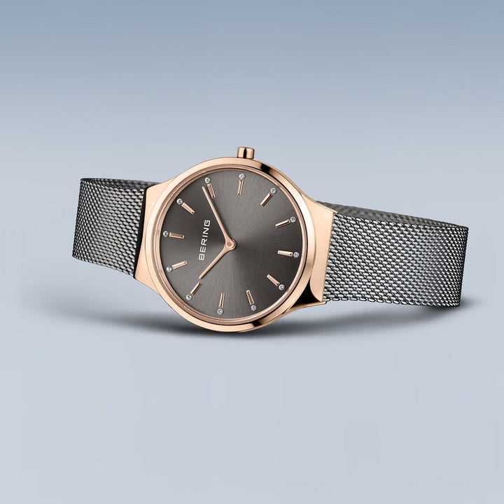 Het Bering Dameshorloge (Product 12131-369-GWP) heeft een roségouden lunette en een grijze wijzerplaat met minimalistische markeringen. Het heeft een fijne zilveren Milanees mesh band. Dit quartz-uurwerk is elegant geplaatst tegen een vloeiende blauwe achtergrond.