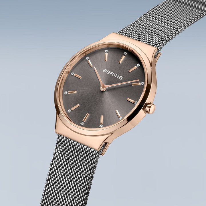 De Bering 12131-369-GWP Dameshorloge toont een roségouden horloge met een grijze sunburst wijzerplaat, zilveren indexen en quartz uurwerk. Het heeft een minimalistische slanke Milanees band, met Bering elegant weergegeven op de wijzerplaat tegen een gradiënt van lichtblauwe tinten.