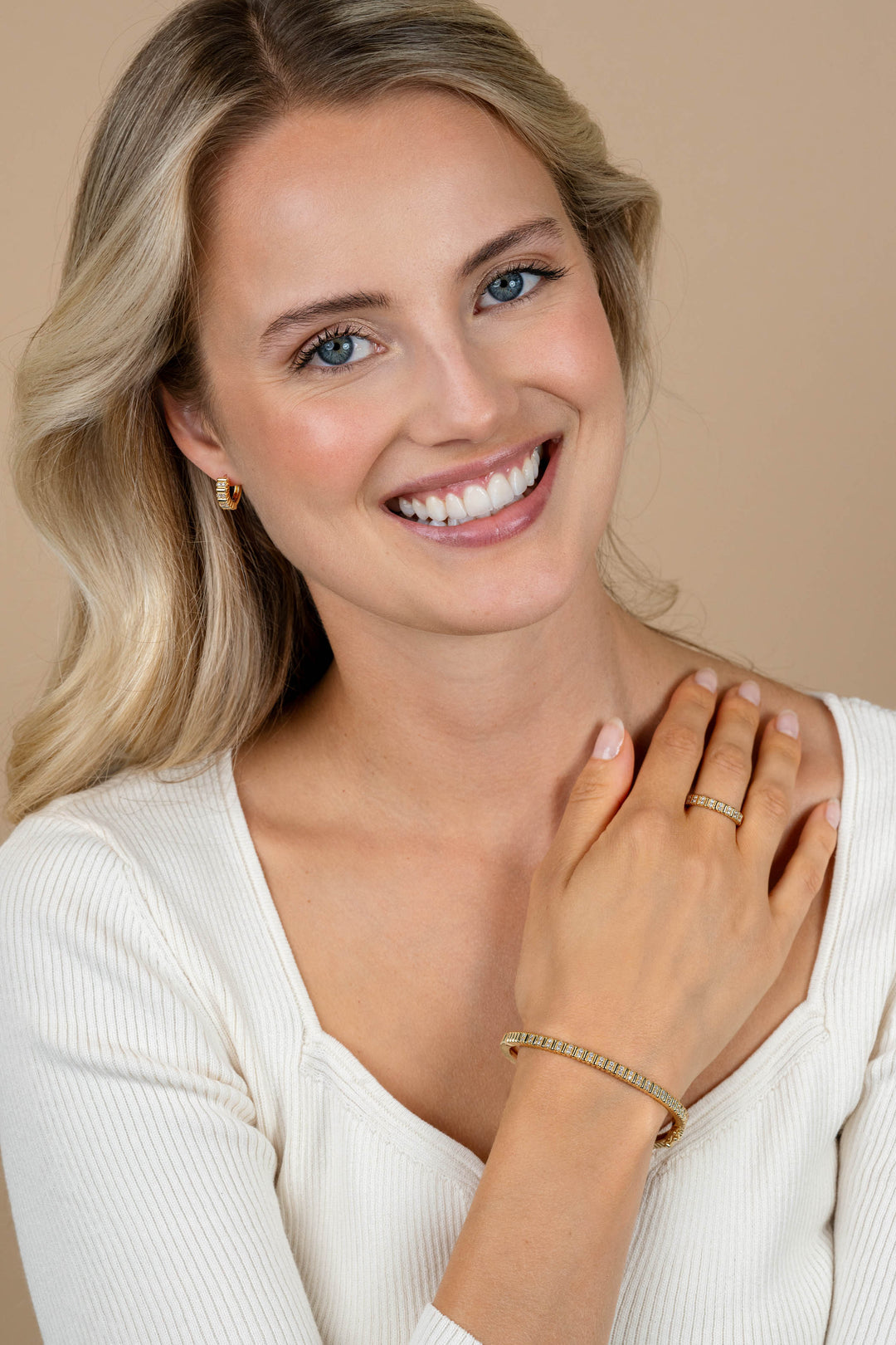 Een lachende blonde vrouw in een wit topje showt de Zinzi Ring Zilver ZIR2686Y van Zinzi, samen met bijpassende tweekleurige gouden sieraden, terwijl ze haar armen over elkaar slaat en naar de camera kijkt tegen een beige achtergrond.