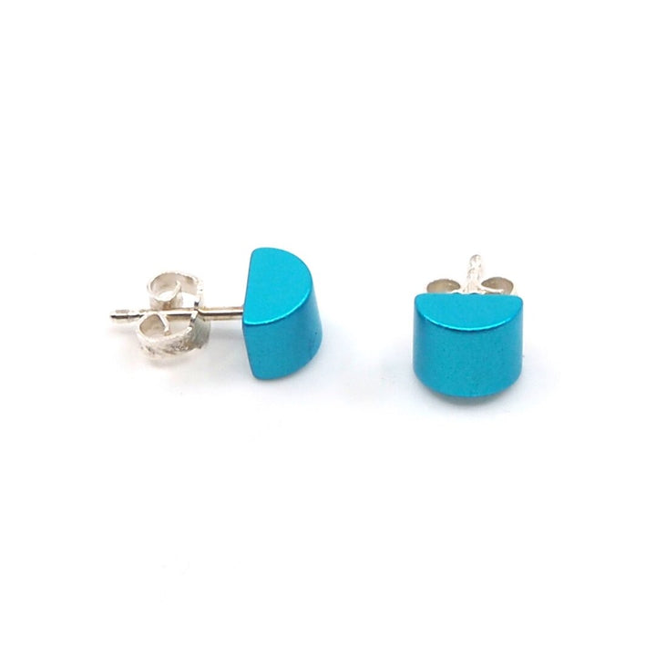 Twee kleine, turquoise halve cilinder oorbellen van Otra Cosa, gemaakt van geanodiseerd aluminium met zilveren staafjes en achterkantjes, afgebeeld op een witte achtergrond, vangen de elegantie van OTRACOSA oorbellen.