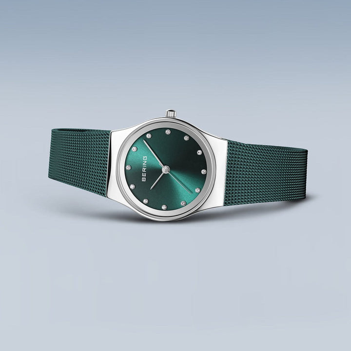 Het Bering Dameshorloge, model 12927-808, heeft een groene wijzerplaat met puntmarkeringen en het BERING-logo. De zilveren kast is gecombineerd met een getextureerd Milanees bandje, allemaal op een grijze achtergrond, wat de tijdloze Quartz-uurwerkprecisie belichaamt.