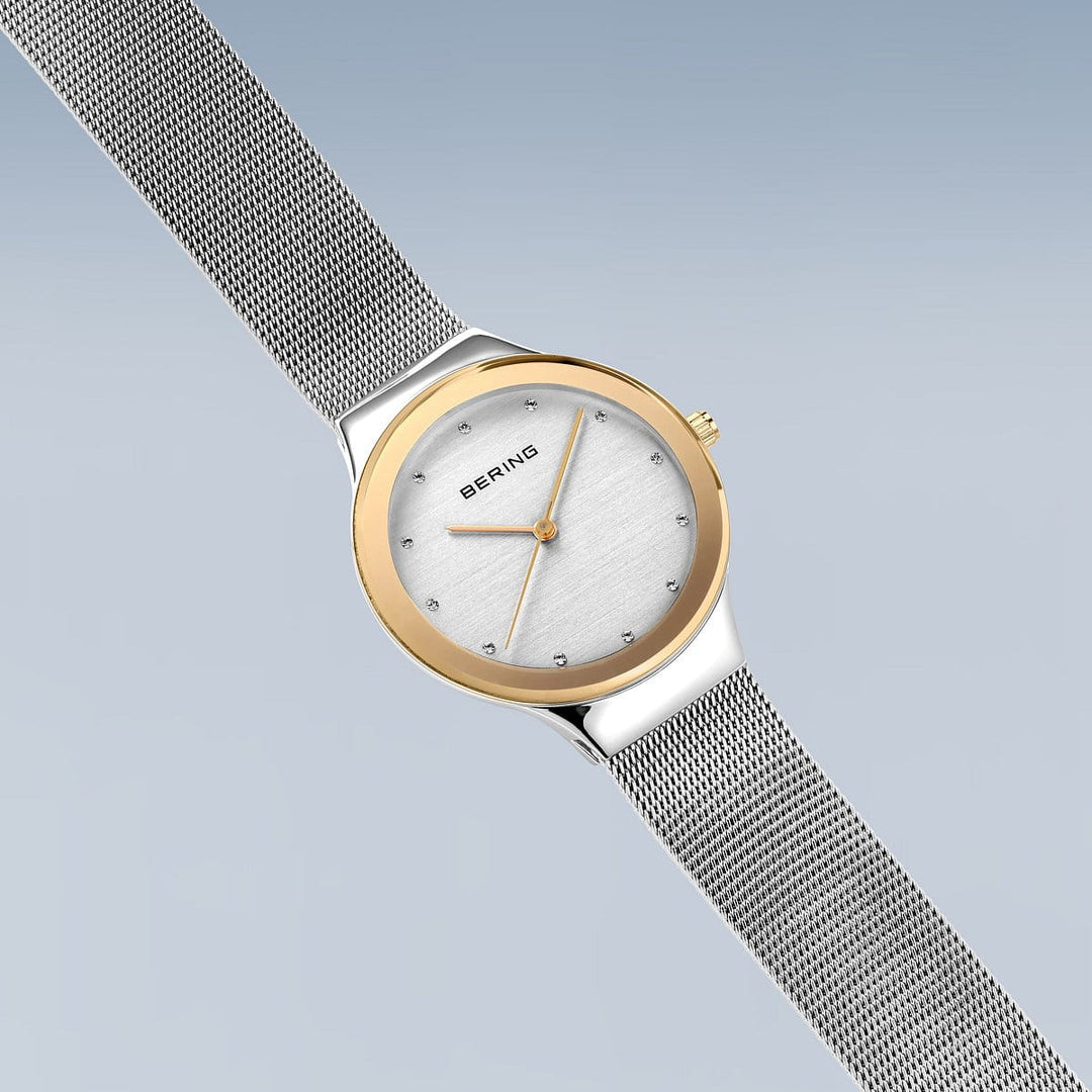 Het Bering 12934-010 horloge heeft een tijdloos design met een ronde wijzerplaat, gouden lunette en mesh zilveren band. Het heeft een zilverkleurige wijzerplaat met gouden wijzers, kleine uurmarkeringen en sluit de roestvrijstalen kast naadloos aan op de band tegen een lichte gradiëntachtergrond.