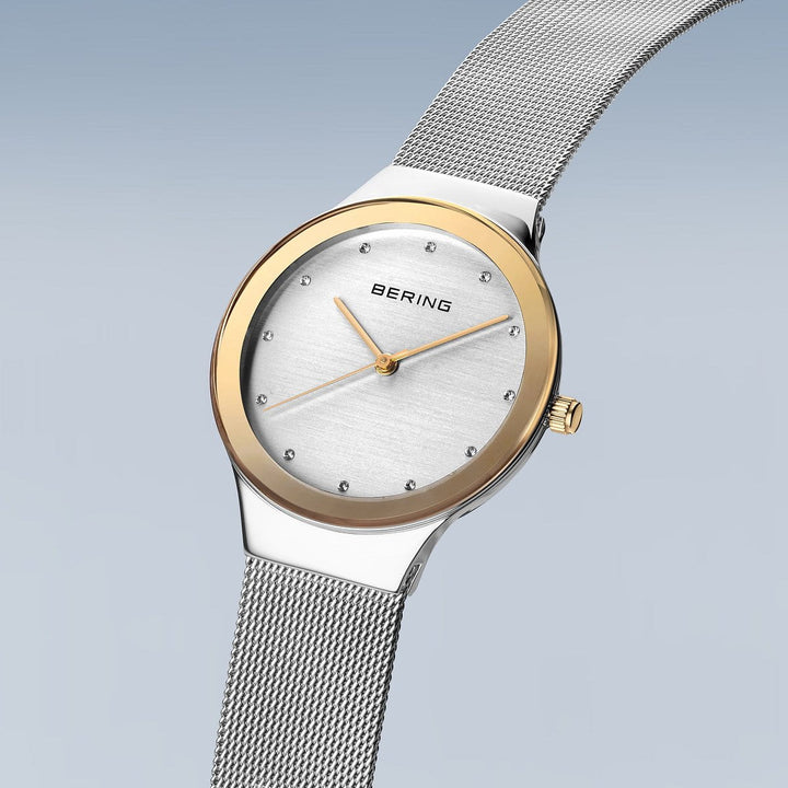 De Bering 12934-010 is een strak dameshorloge met een tijdloos design, voorzien van een roestvrijstalen kast en zilveren mesh band. Het heeft een witte wijzerplaat met gouden lunette, zilveren en uurmarkeringen. De naam Bering valt op op de lichtblauwe gradient achtergrond.