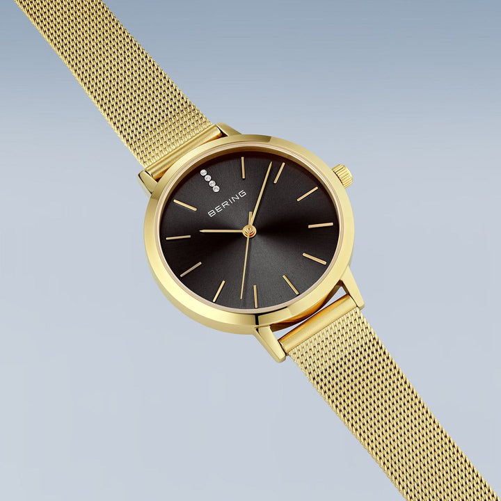 Het Bering dames horloge 13434-332 heeft een zwarte wijzerplaat met gouden minimalistische uurmarkeringen, saffierglas en een goudkleurige Milanees band. BERING staat op de wijzerplaat, gepresenteerd tegen een lichtblauwe achtergrond.