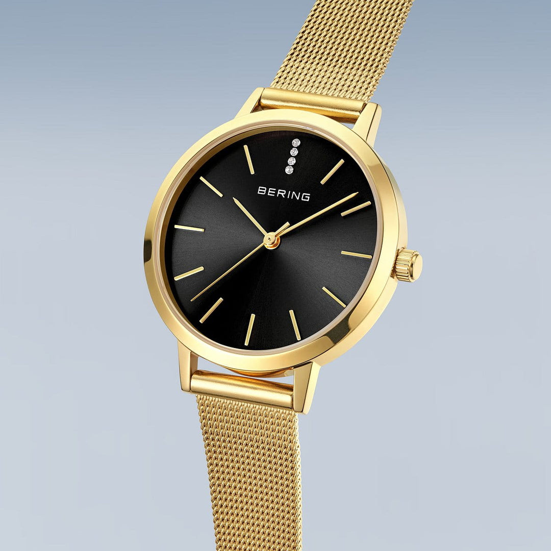 De Bering dames horloge zwarte wijzerplaat 13434-332 heeft een gouden kast, zwarte wijzerplaat met minimalistische markers, drie kristallen op 12 uur, een goudkleurige Milanees band en saffierglas op een lichtverloop achtergrond.