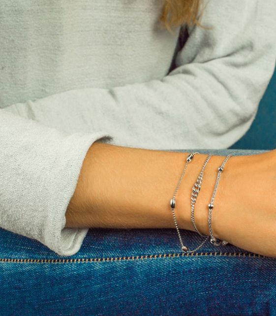 Close-up van de arm van een persoon op een blauwe spijkerbroek, versierd met drie delicate zilveren armbanden, waaronder de prachtige Zinzi Armband Zilver Zia1128. Ze dragen een lichtgrijze top met lange mouwen, waardoor de sieraden en de textuur van de stof worden benadrukt.