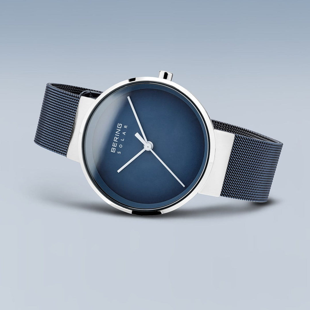Het Bering Unisex Horloge 14339-307 is een minimalistisch horloge met een donkerblauwe wijzerplaat en zilveren kast. Het heeft dunne witte wijzers, een blauwe mesh band en is voorzien van Bering en Solar gravures op de gradient achtergrond, wat een elegant accessoire in moderne stijl creëert.