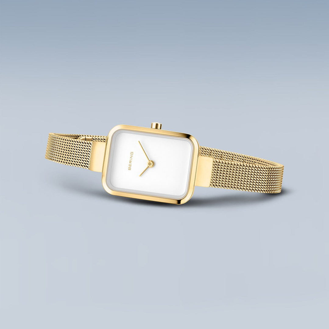 Het Bering horloge 14520-334 wordt getoond tegen een lichtblauwe achtergrond. Dit goudkleurige model heeft een vierkante witte wijzerplaat, minimalistisch design, mesh metalen band en gouden wijzers en roestvrijstalen kast.
