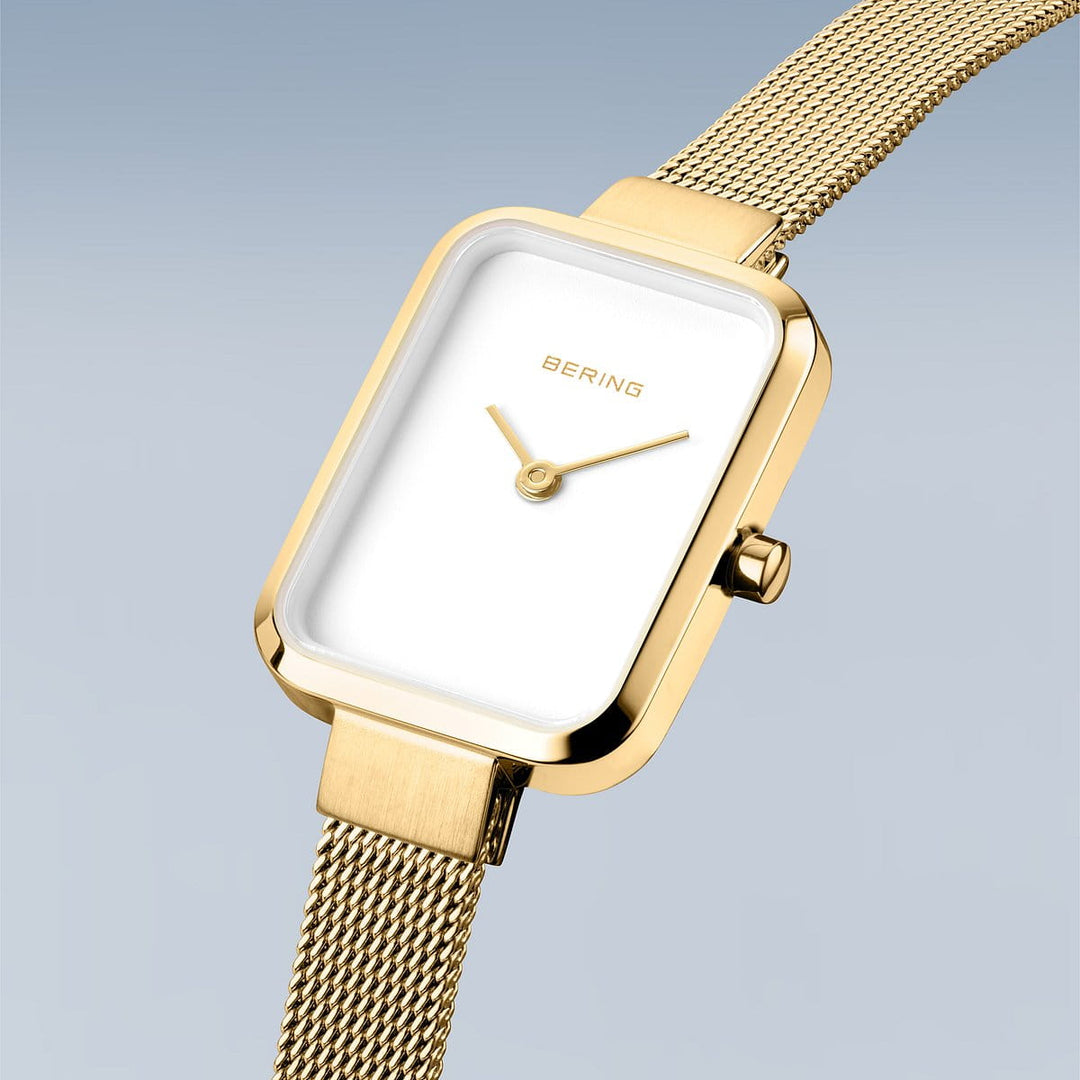 De Bering 14520-334 dameshorloge heeft een rechthoekige witte wijzerplaat met een minimalistisch ontwerp, geen uurmarkeringen en een goudkleurige mesh band, afgebeeld tegen een lichtblauwe achtergrond.