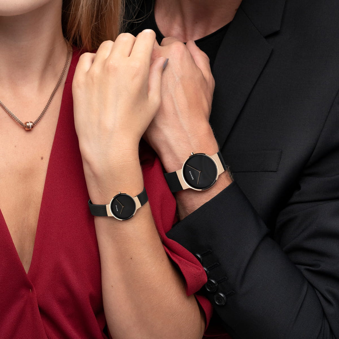 Een vrouw in een rode jurk en een man in een zwart pak kruisen hun armen, beiden dragen ze strakke zwarte horloges met minimalistische ontwerpen. Haar horloge is een Bering Dames Horloge 14531-166 met een quartz uurwerk, kleiner formaat en roestvrijstalen kast, perfect passend bij haar ronde hangerketting.