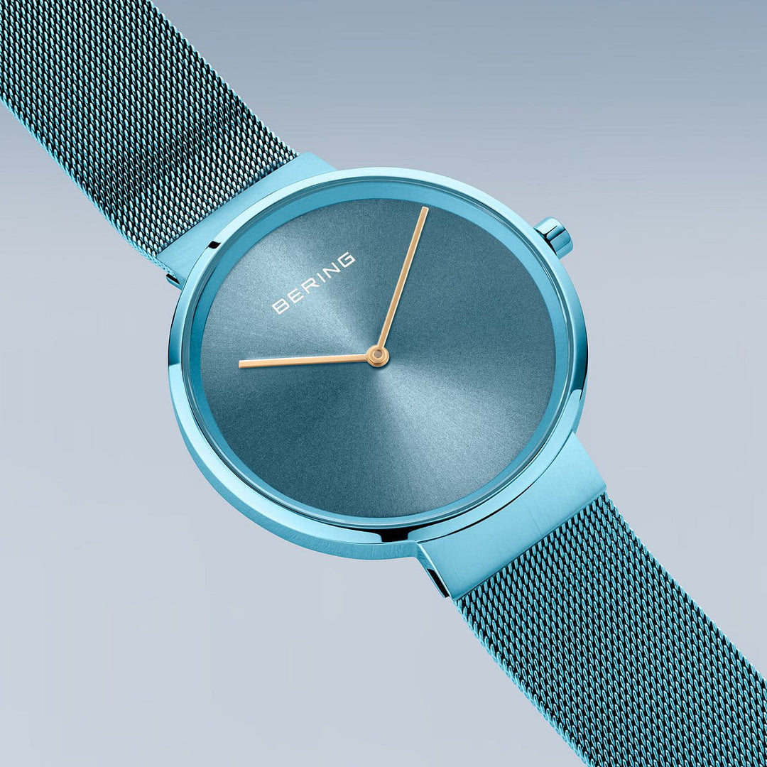 De Bering 14539-388, een strak turquoise horloge met een mesh band, staat op een gradiëntblauwe achtergrond. De minimalistische wijzerplaat heeft gouden uur- en minutenwijzers onder Saffierglas. BERING staat op de 12-uurspositie en toont Quartz-uurwerk precisie.