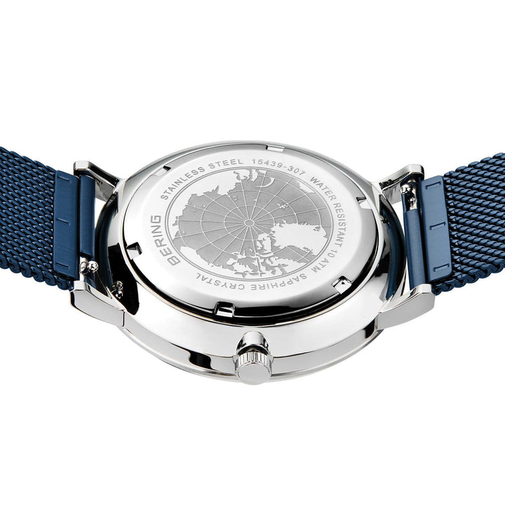 De afbeelding toont een Bering 15439-307 Herenhorloge, een zilveren polshorloge met een blauwe meshband. De achterkant toont een gegraveerde wereldkaart met labels voor roestvrij staal, waterbestendigheid, saffierglas en het modelnummer van dit solar-uurwerk.