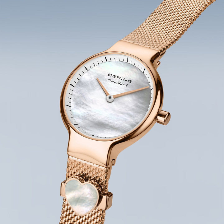 Het Bering horloge model 15527-364-GWP heeft een gouden mesh band, witte parelmoer wijzerplaat met zwarte wijzers en een hartvormig accent. Het heeft Saffierglas voor duurzaamheid en een elegante blauwgrijze gradiënt achtergrond.