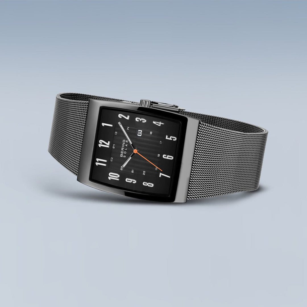 Het Bering 16433-377 polshorloge heeft een strak rechthoekig ontwerp met een zwarte wijzerplaat, witte cijfers en een oranje secondewijzer. Dit roestvrijstalen horloge heeft een zilveren meshband en minimalistische stijl, op zonne-energie tegen een gradiënt blauwgrijze achtergrond.