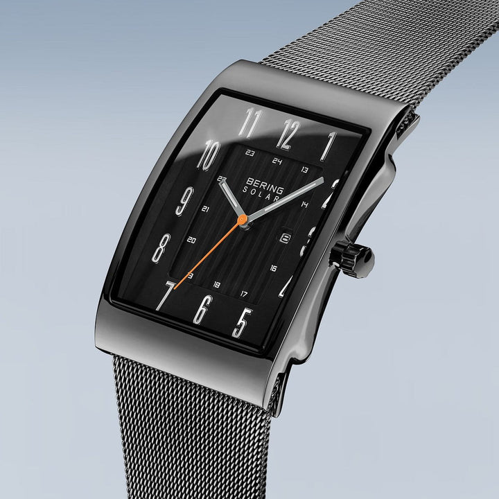 De Bering 16433-377 is een zilverkleurig rechthoekig horloge met een zwarte wijzerplaat, witte cijfers en een oranje secondewijzer. Het roestvrijstalen horloge heeft een meshband en is voorzien van Bering Solar op de wijzerplaat. De achtergrond vervaagt van lichtblauw naar grijs, wat de zonne-energie horlogefunctie benadrukt.