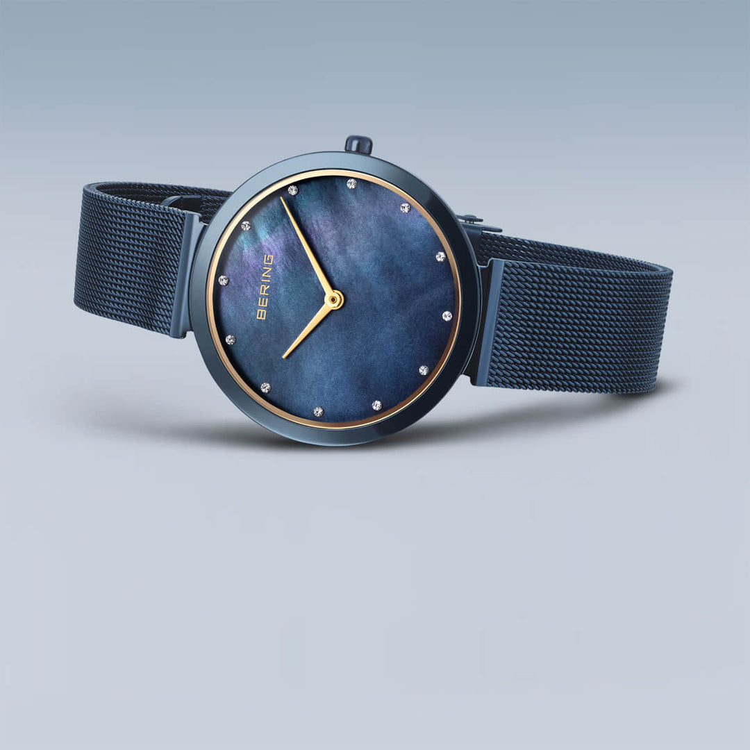 Een close-up van een strak Bering dameshorloge, model 18132-399, toont de Milanees band en het minimalistische design met ronde uurmarkeringen en twee wijzers op een blauwe wijzerplaat. Het merk Bering is prominent weergegeven op de subtiel verlopende wijzerplaat.