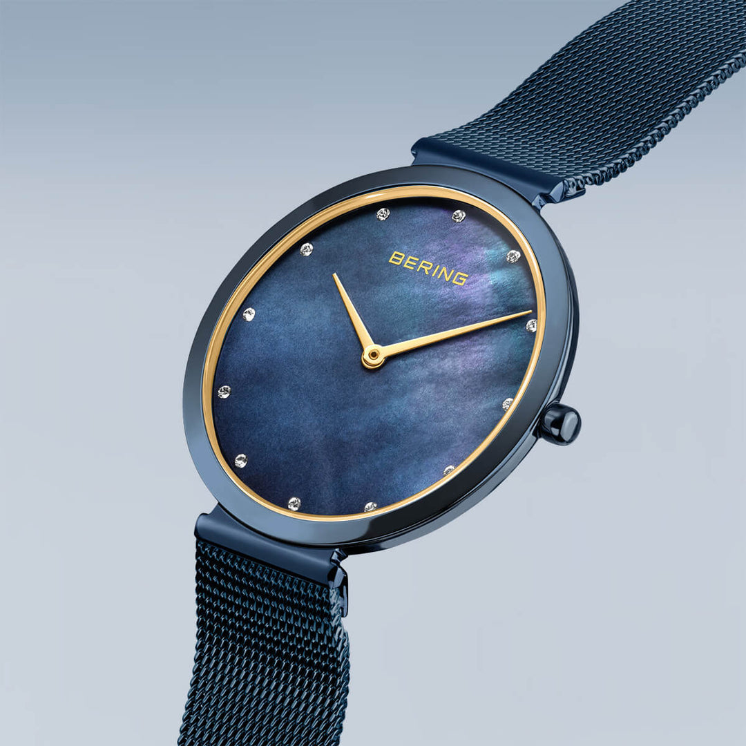 Een strak Bering dameshorloge met een donkere Milanees band, met een glanzende ronde wijzerplaat met een blauw-zwarte gradiënt en kleine ronde markeringen. De minimalistische wijzers complementeren het elegante ontwerp met BERING tegen een lichtgrijze achtergrond. Product: bering dameshorloge blauwe dial - 18132-399.