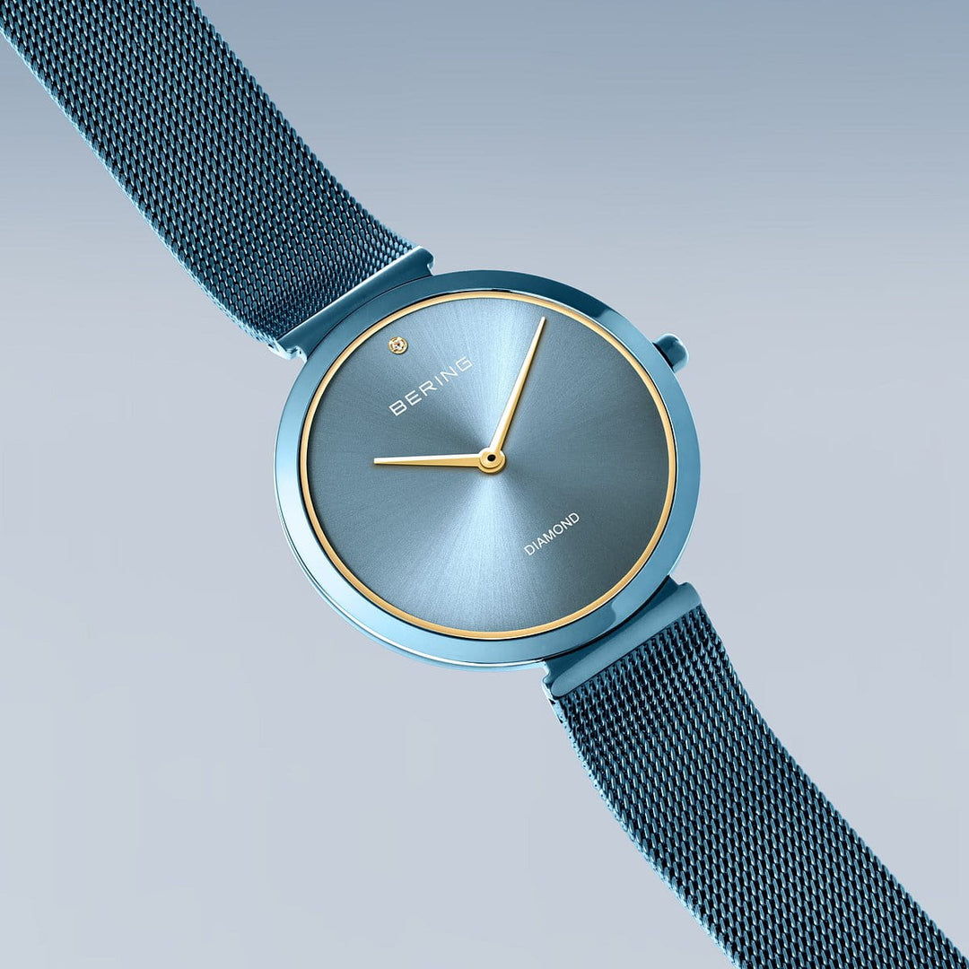 De Bering dames horloge blauwe wijzerplaat - 18132-Charity1 heeft een minimalistische blauwe wijzerplaat, goudomrande kast, eenvoudige gouden wijzers, een diamant op 12 uur en een blauwe RVS mesh horlogeband, weergegeven op een lichtblauwe achtergrond.