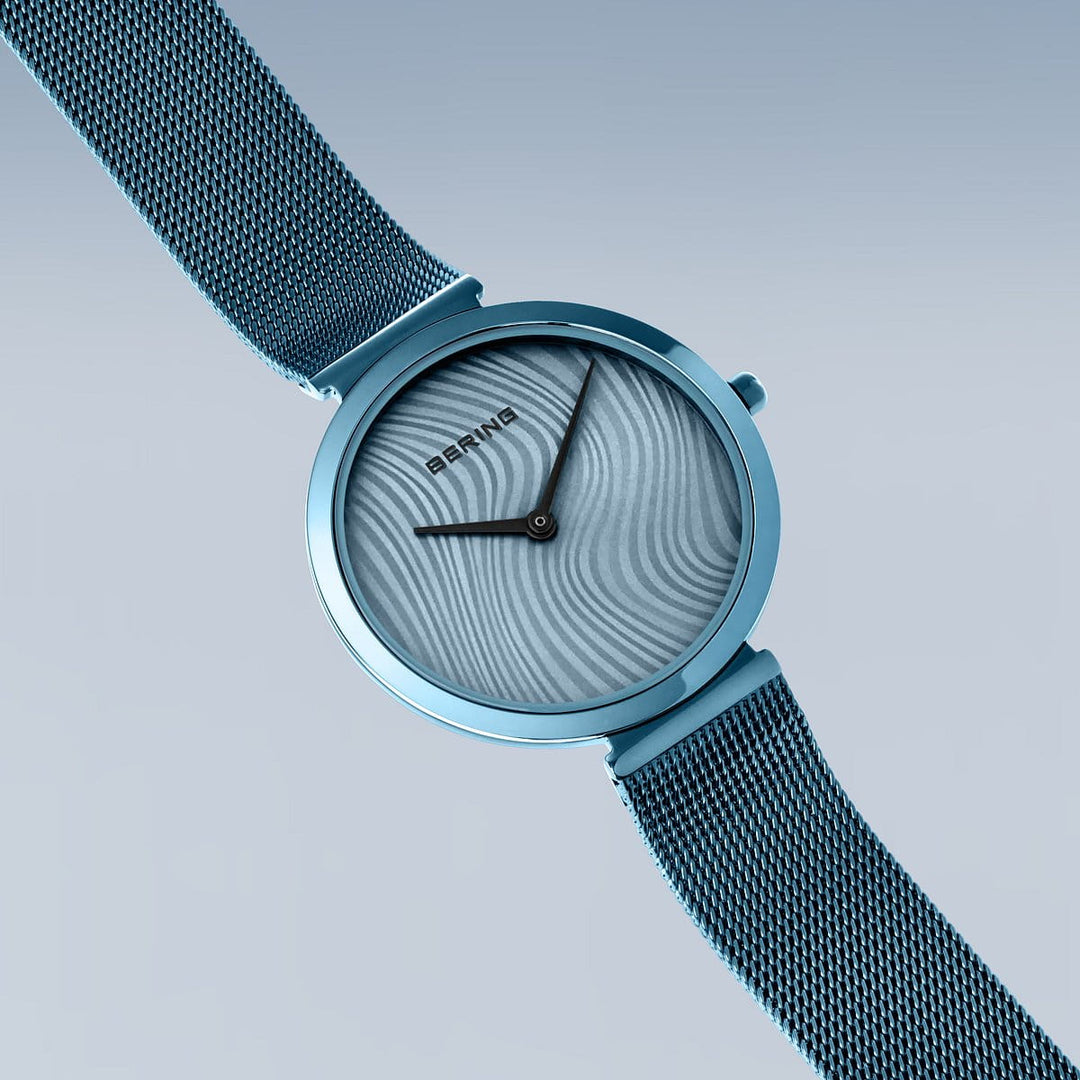 Het Bering dames horloge 18132-Charity2 heeft een blauwe wijzerplaat met golvende structuur en zwarte wijzers, een metalen horlogeband en saffierglas. Weergegeven op een lichtblauwe gradiënt achtergrond is het Bering logo zichtbaar op de wijzerplaat.