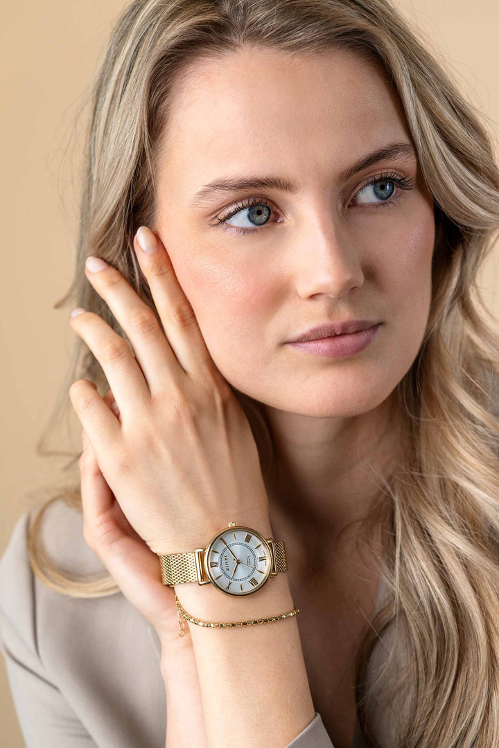 Een vrouw met lang blond haar en een beige topje kijkt peinzend opzij. Om haar linkerpols draagt ze het Zinzi Geelgoudkleurig Horloge Gold Plated Ziw2033 van Zinzi.