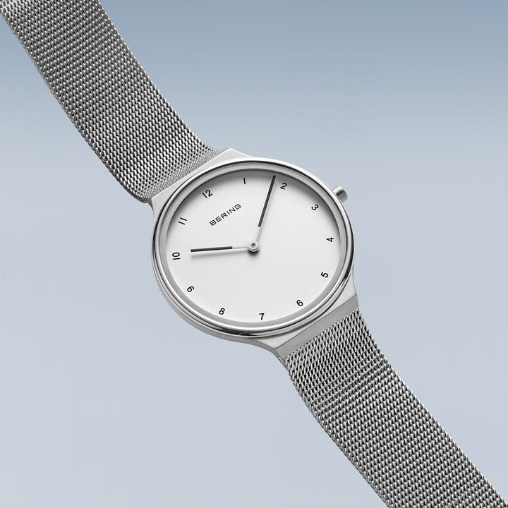 Het Bering dameshorloge model 18434-004 heeft een minimalistisch design met een gradiënt blauwe achtergrond, witte wijzerplaat, eenvoudige zwarte uurmarkeringen en dunne wijzers, allemaal beschermd door duurzaam saffierglas. Het bevat een zilveren mesh band en is van het bekende merk Bering.