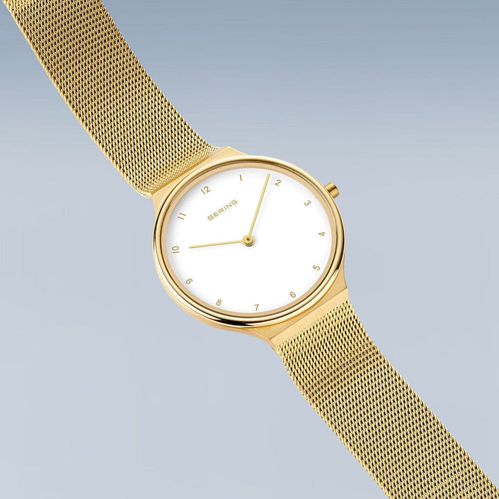 Het Bering dameshorloge goud - 18434-334 heeft een minimalistisch design met een witte wijzerplaat, gouden uurmarkeringen en een meshband, tegen een zachte blauwgrijze achtergrond.