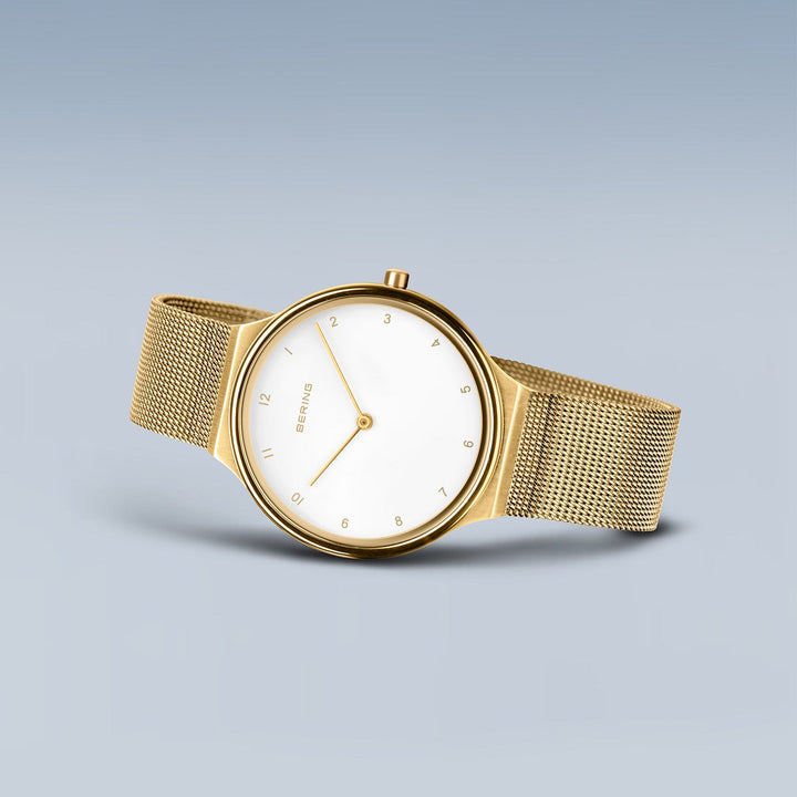 Het Bering dames horloge goud - 18434-334 heeft een minimalistisch goud design met een witte wijzerplaat, gouden mesh band, zwarte uurmarkeringen en twee wijzers. Het is geplaatst tegen een gradiënt blauwe achtergrond.