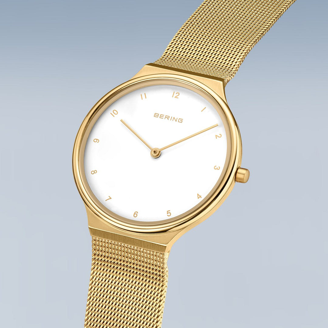 Het Bering dames horloge goud - 18434-334 is een minimalistisch gouden polshorloge met een witte wijzerplaat, gouden uurmarkeringen en delicate gouden wijzers, aangevuld met een slank gouden meshbandje. Het staat op een zachtblauwe achtergrond met kleurverloop.