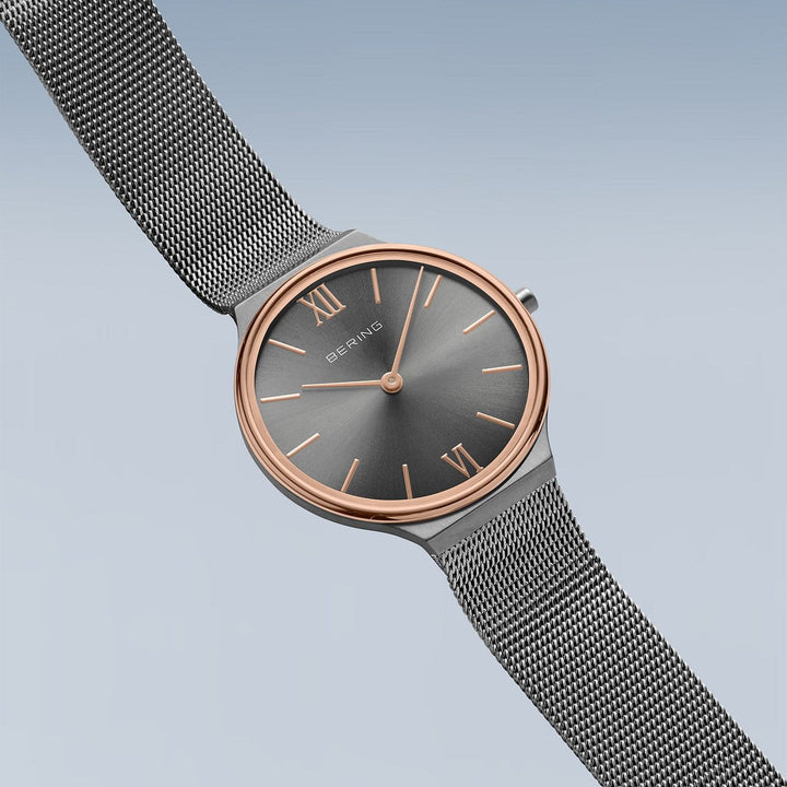Het Bering dameshorloge 18434-369 heeft een strak, minimalistisch design met een metalen mesh band, een donkergrijze wijzerplaat met een roségouden accent, een quartz uurwerk beschermd door saffierglas, Romeinse cijfers op 12 en 6 uur en eenvoudige uurmarkeringen op een zachtblauwe achtergrond.