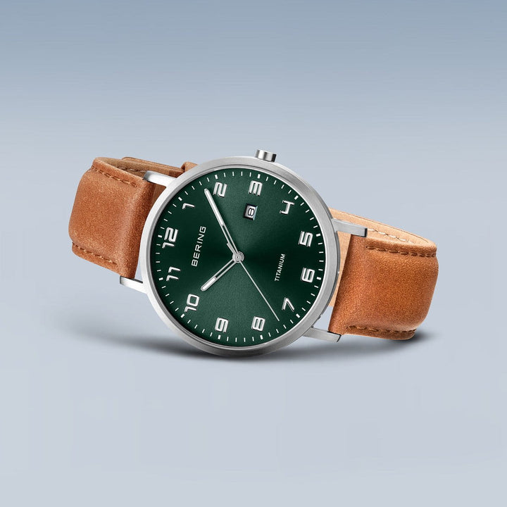 Het Bering herenhorloge 18640-568 heeft een strak design met een groene wijzerplaat, witte uurmarkeringen en een datumvenster. Het is omhuld met titanium en heeft saffierglas. Dit elegante exemplaar wordt gecomplementeerd door een bruine leren band tegen een lichtgrijze achtergrond.