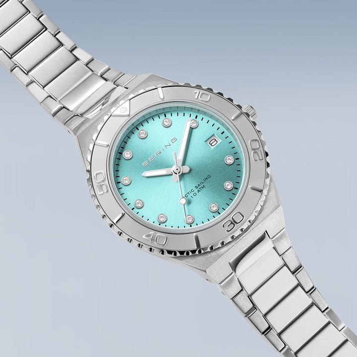 Het Bering dameshorloge (model 18936-707) heeft een turquoise wijzerplaat met diamanten uurmarkeringen, de merknaam bovenaan en SWISS PARTS eronder. Het heeft een waterdichtheid van 10 ATM, een metalen schakelband, lichtblauwe genummerde lunette en duurzaam saffierglas.