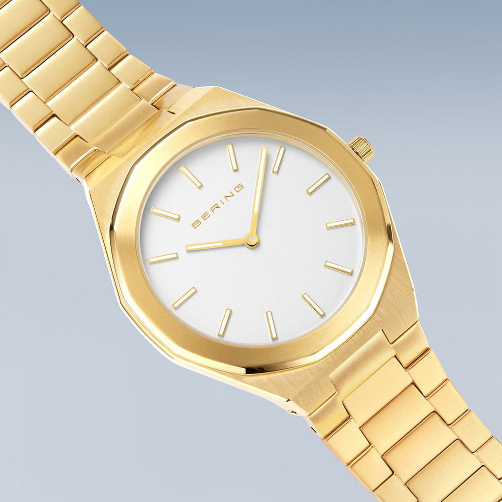 Het Bering herenhorloge 19641-730 heeft een gouden geometrische kast en goudkleurige roestvrijstalen band tegen een lichtblauwe achtergrond. Het heeft een witte wijzerplaat met gouden markeringen en wijzers, waarbij de merknaam prominent op de wijzerplaat staat.