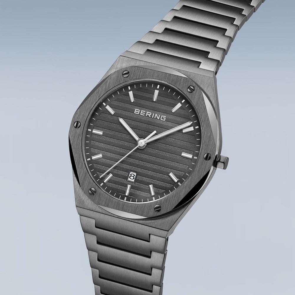 Een strak Bering herenhorloge met een roestvrijstalen kast en metalen band heeft een hexagonale lunette, slanke witte uurmarkeringen en wijzers op een getextureerde zwarte wijzerplaat onder duurzaam saffierglas. Het Bering-logo is prominent aanwezig en er is een klein datumvenster op 6 uur. Model: 19742-777.