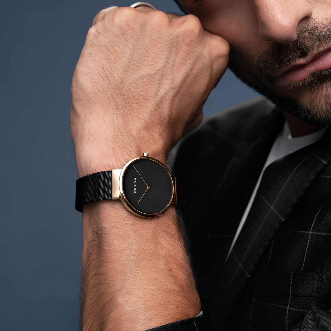 Een persoon draagt het Bering 14539-166 horloge, een strak zwart model met een gouden rand en een minimalistisch design. Het quartz-uurwerk siert hun linker pols en complementeert hun zwart geblokte pak tegen een effen blauwgrijze achtergrond die het gezicht gedeeltelijk omlijst.
