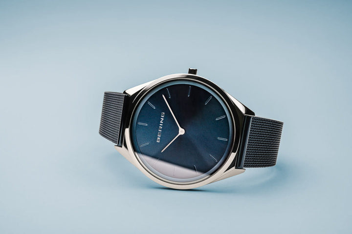 Het Bering 17039-307 unisex horloge heeft een strakke donkerblauwe wijzerplaat, zilveren kast, minimalistische uurmarkeringen, saffierglas en een fijn geweven zwarte band. Dit quartz horloge, weergegeven op een lichtblauw oppervlak, belichaamt hedendaagse stijl en elegantie.