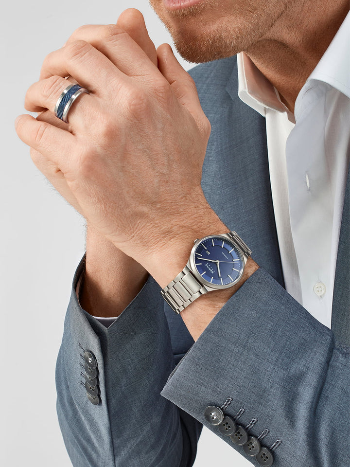 Een persoon in een grijs pak met bijpassende mouwknopen en een wit overhemd vouwt zijn handen, waarbij een zilveren ring zichtbaar is. Om zijn linker pols draagt hij een Bering horloge, model 15239-777, met een blauwe wijzerplaat en een titanium band.