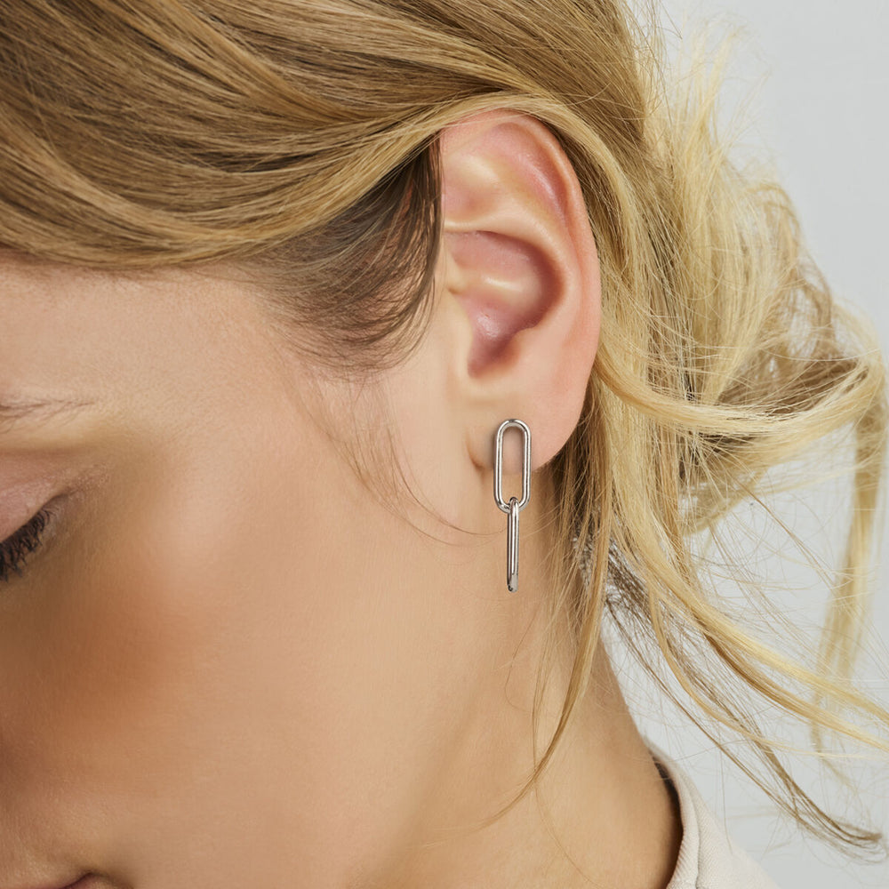 Een close-up van het oor van een vrouw met Van der Kooij Collectie oorhangers paperclip zilver gerhodineerd, voorzien van geometrische druppelvormen met rechthoekige en verticale streepjes. Haar losjes gestylde blonde haar bedekt gedeeltelijk deze elegante dames oorbellen.