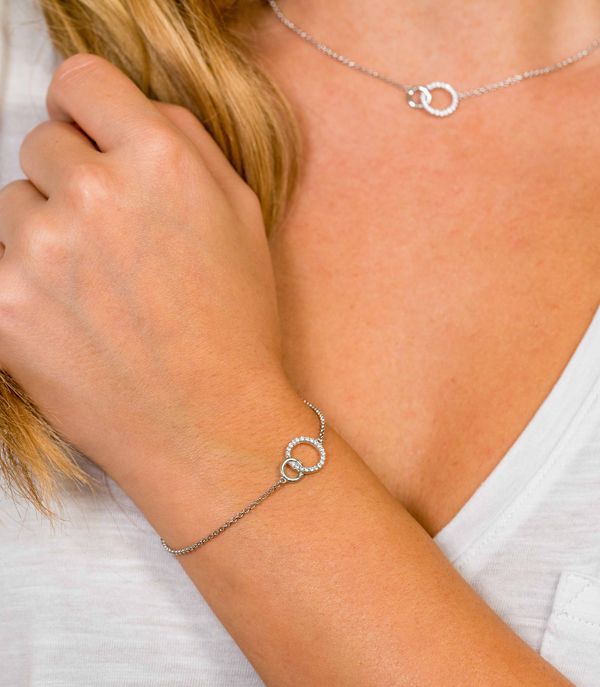Een persoon met lichtbruin haar in een wit shirt draagt de Zinzi Armband Zilver Zia1782 en een bijpassende zilveren ketting, beide met in elkaar grijpende ringen met kristallen. Hun hand rust bij hun sleutelbeen.
