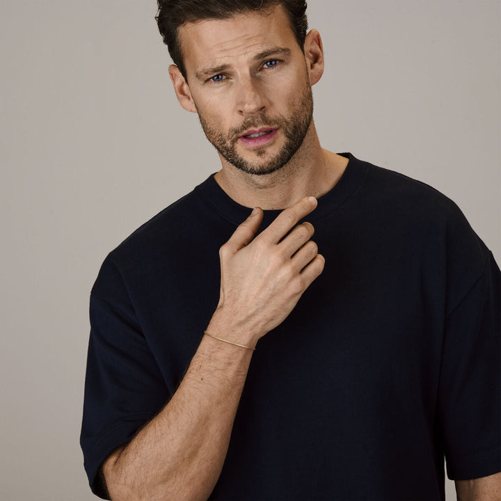 Een man met kort bruin haar en een baard draagt een donker navy T-shirt en de Van der Kooij Collectie Gouden armband heren slang rond 1,2 mm 14K, kijkt serieus naar de camera terwijl hij zijn nek aanraakt tegen een neutrale achtergrond.