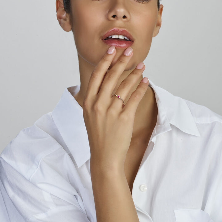 Een vrouw in een wit overhemd strijkt zachtjes over haar gezicht en benadrukt een Van der Kooij Collectie ring met geboortesteen robijn juli 14K geelgoud aan haar linkerhand. Haar lippen zijn lichtjes gespleten en haar nagels zijn netjes gemanicuurd.