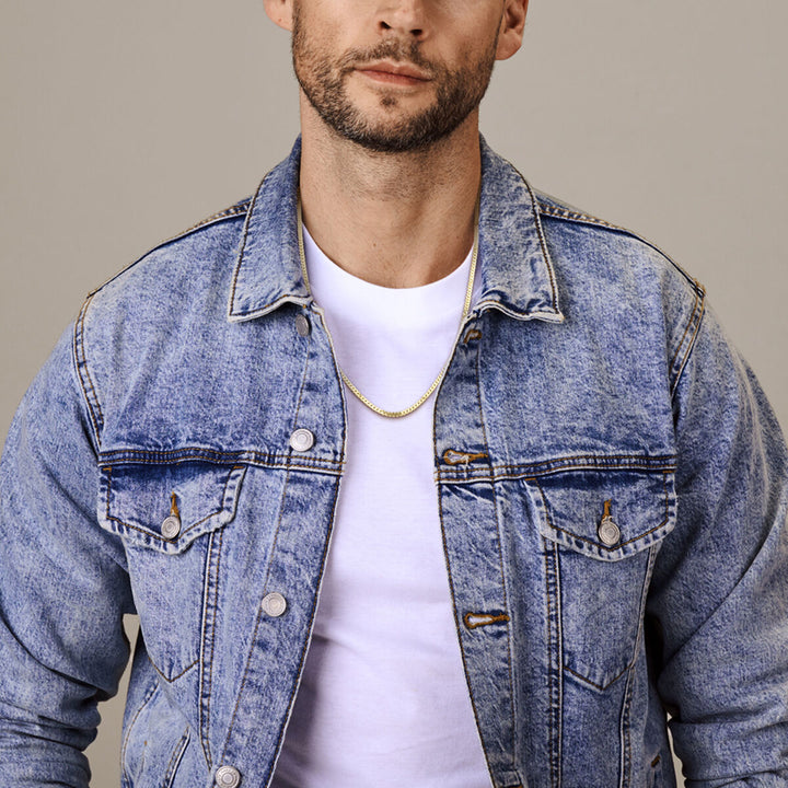 Een man draagt een lichtblauw spijkerjack over een wit T-shirt en de Van der Kooij Collectie Gouden ketting heren - gourmette 4-zijdes geslepen 2,7 mm massief 14K, afgebeeld van schouders tot middenborst tegen een neutrale achtergrond.