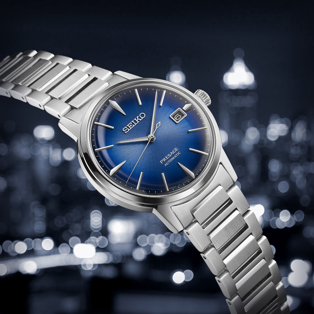 Het Seiko Presage Heren Horloge SRPJ13J1, een zilveren automatisch horloge met een roestvrijstalen kast en metalen band, heeft een blauwe wijzerplaat met een datumvenster. Het wordt weergegeven tegen een verlichte wazige stadsgezichtachtergrond.