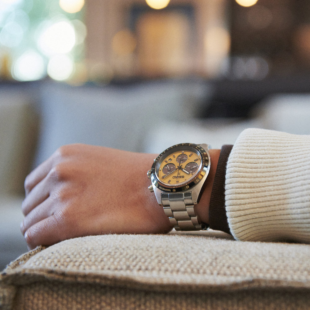 Een close-up toont een pols met een Seiko Prospex heren horloge SSC817P1 met saffierglas, een zilveren metalen band en een gouden wijzerplaat op beige stof. De zacht gefocuste achtergrond hint naar wazige binnenelementen en lichten, terwijl de drager een crèmekleurige trui draagt.